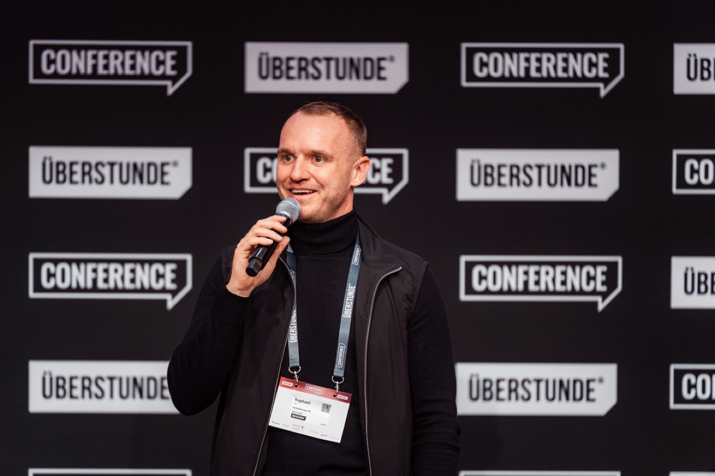 Speaker auf der Bühne mit Namensschild am Lanyard vor Konferenz-Backdrop